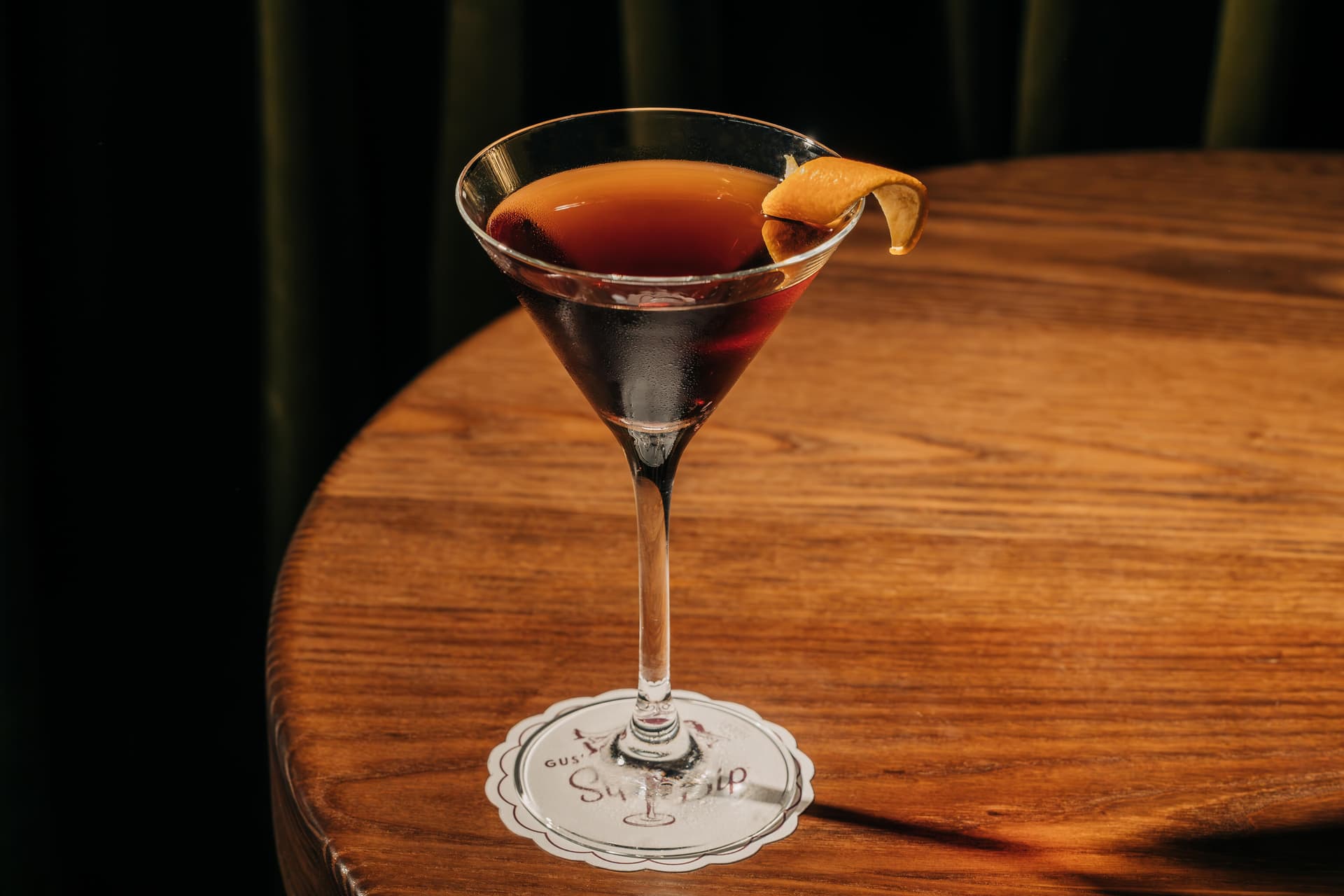 A cocktail glass on a wooden table contains a dark liquid, garnished with a twist of orange peel.