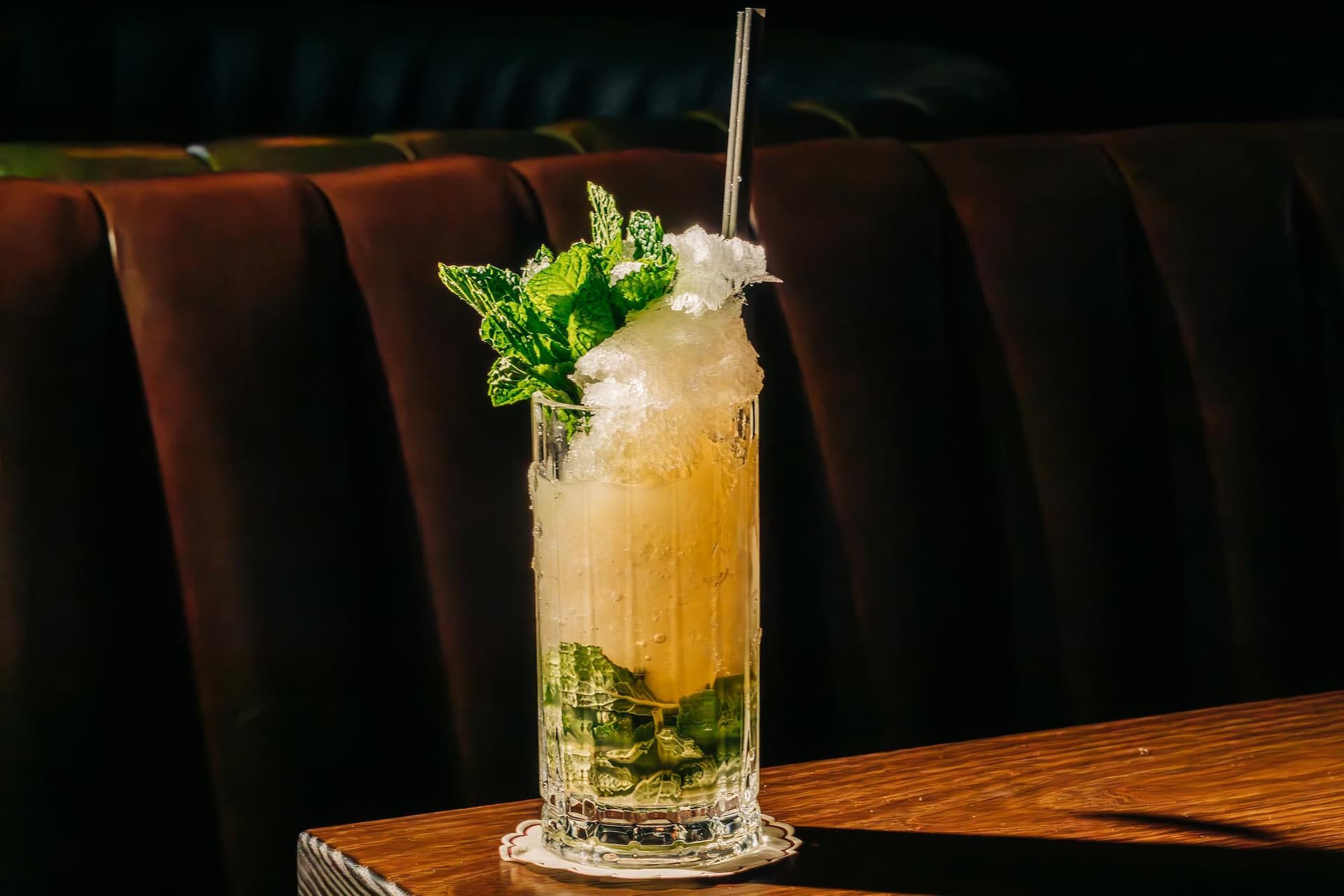 A tall cocktail glass filled with a mint julep, garnished with fresh mint leaves and a straw, placed on a dark wooden table.