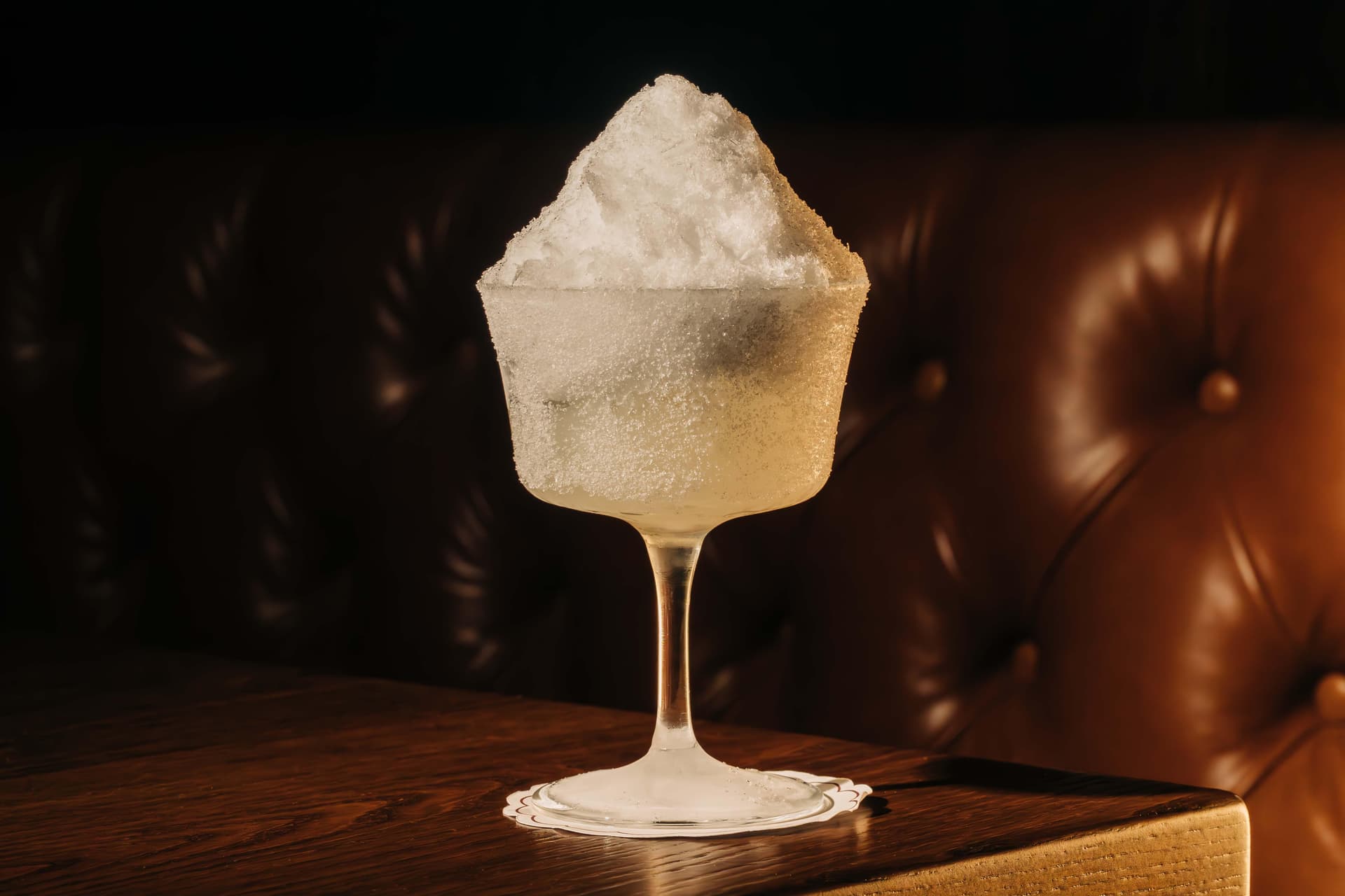 A frosty margarita cocktail with a mound of shaved ice, served in a frosted glass on a wooden table. The background features a leather-upholstered surface.