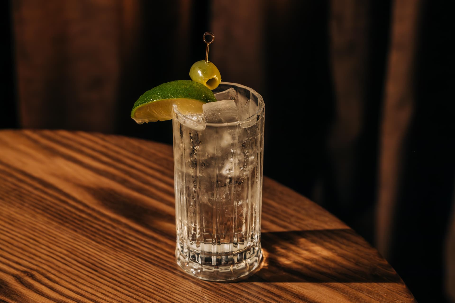 A glass of clear cocktail with ice, garnished with a lime wedge and a green olive on a skewer, placed on a wooden table.