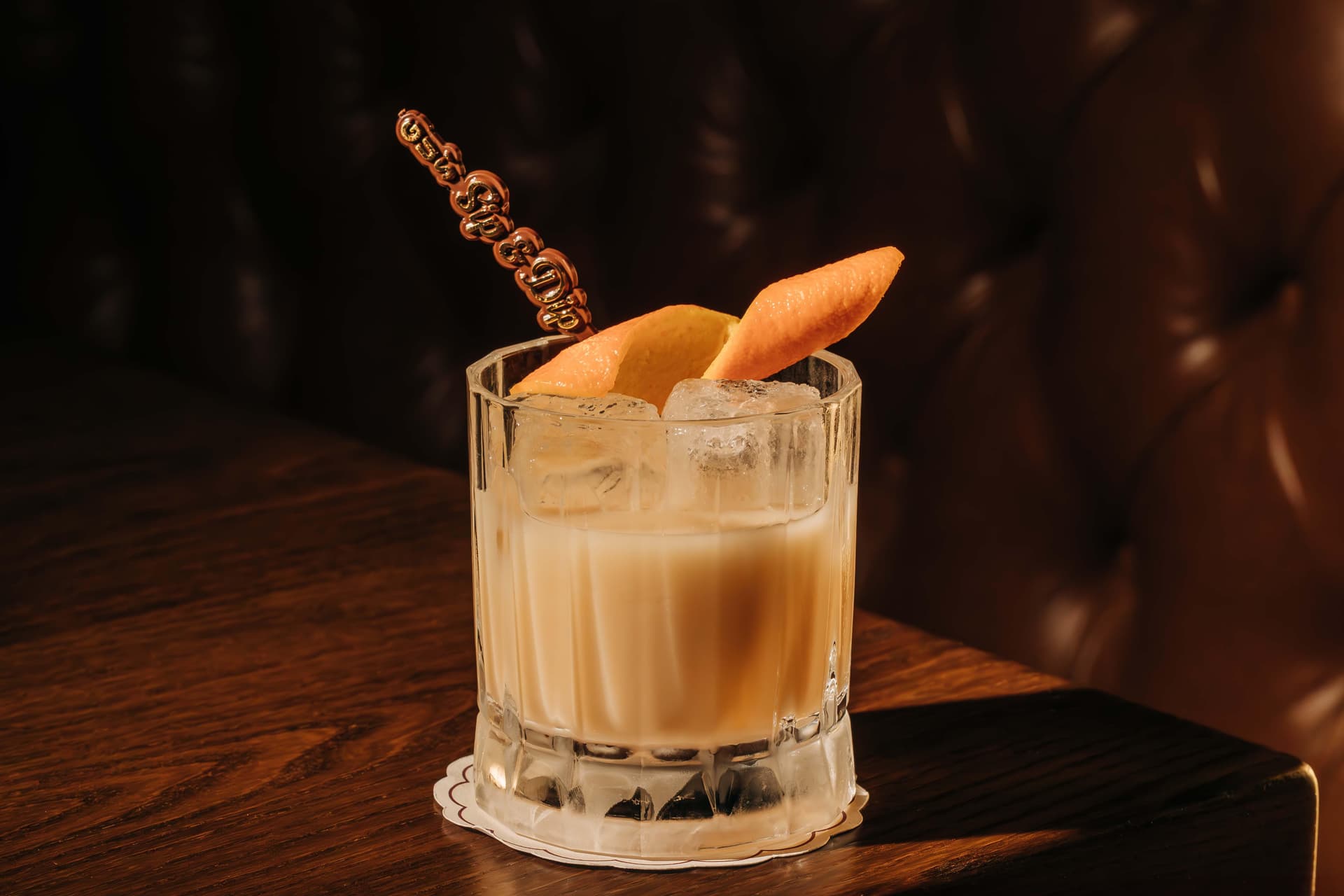 A creamy cocktail garnished with citrus peel sits on a wooden table, backdropped by a tufted leather booth.