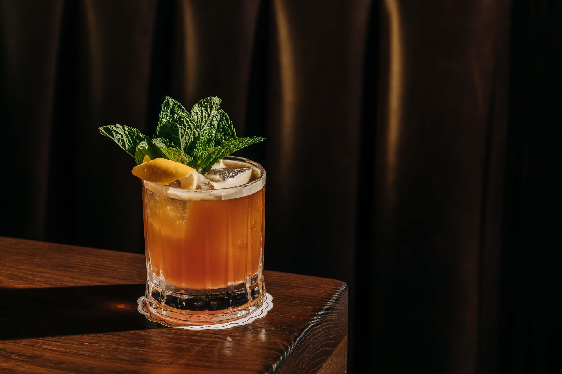 A glass of iced cocktail garnished with mint leaves and a lemon slice, placed on a wooden surface against a dark background.