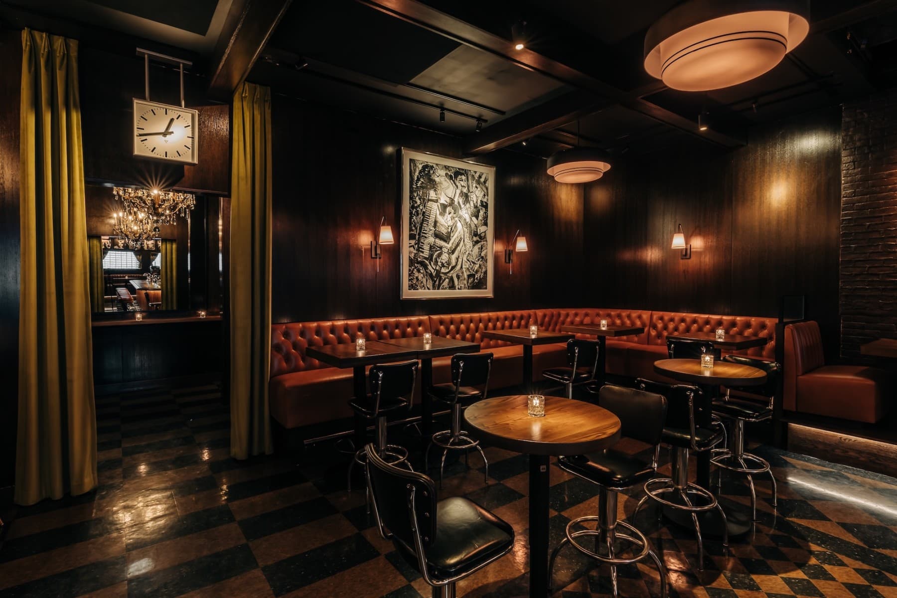 Interior of a dimly lit restaurant with an elegant bar area, featuring reddish-orange leather booths, dark wooden tables with matching chairs, and a large intricate art piece on the wall. The floor is glossy with a checkered pattern and there are curtain accents beside a doorway leading to another area.