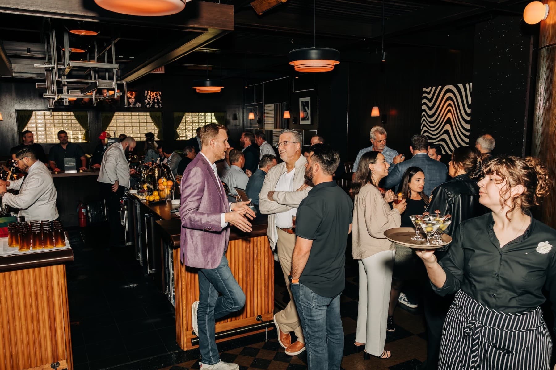 A bustling bar scene with multiple people engaged in conversation and a server carrying drinks.