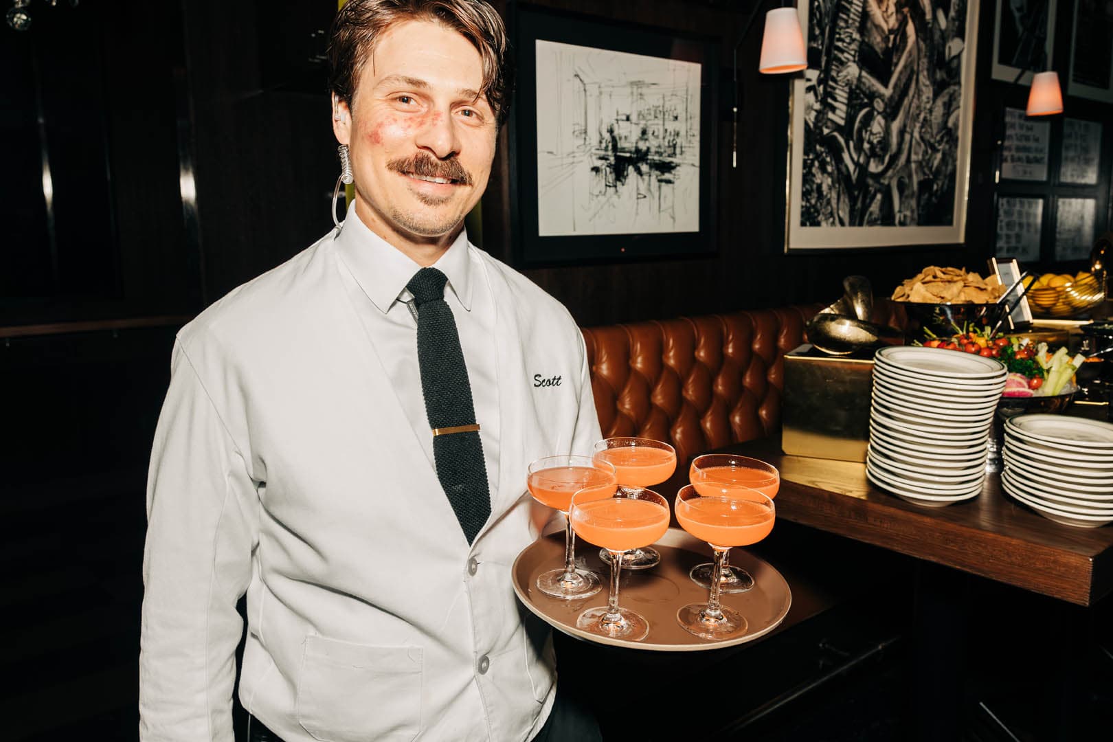 A staff member at Gus' Sip & Dip stands beside a table holding five Naked & Famous cocktails, with plates and a dark, elegant interior in the background.
